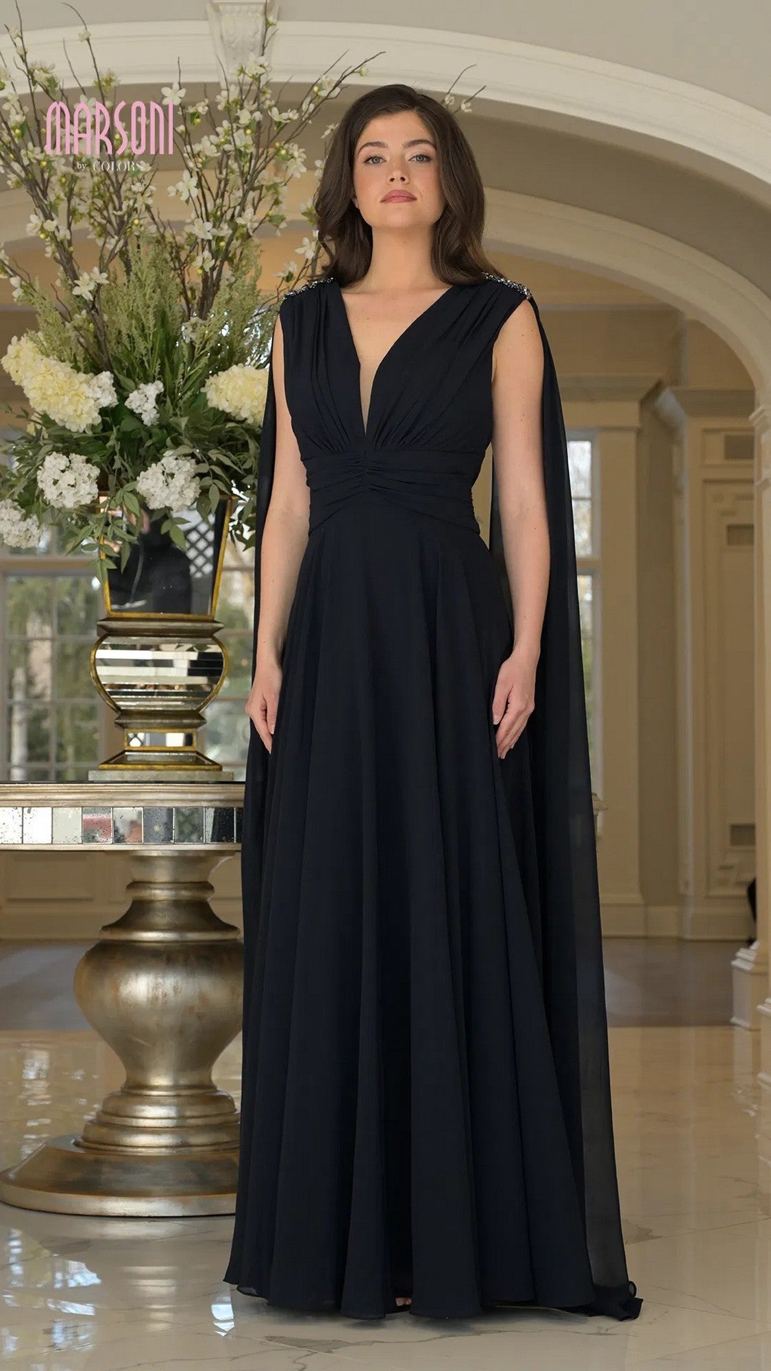 A woman stands indoors in front of a large floral arrangement wearing the Marsoni MV1349 long formal dress in midnight, featuring black chiffon fabric, long cape sleeves, a deep V-neck, and a ruched bodice.